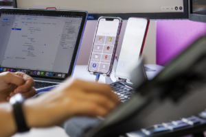 Two mobile phones and a laptop on stands on a desk. In the foreground hand types on a computer keyboard.