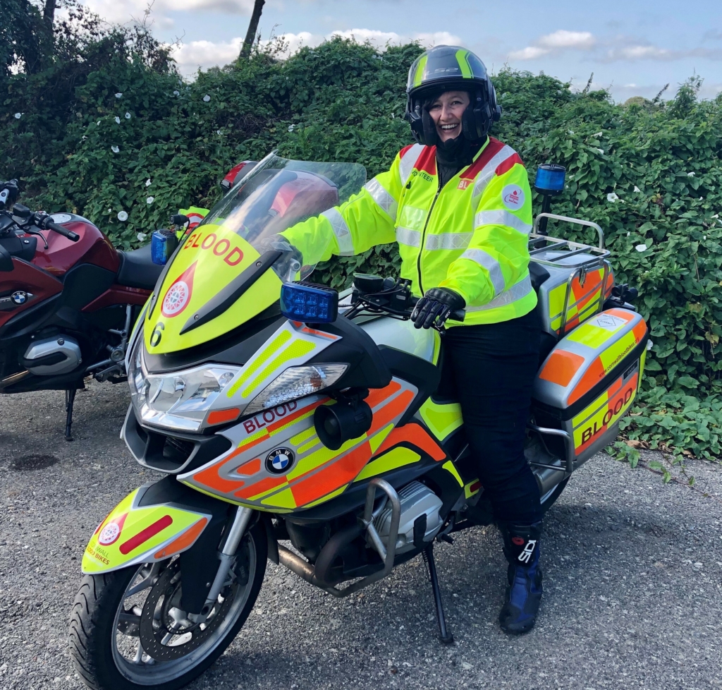 A motorcyclist in helmet and high-vis sits on BMW motorbike with high-vis emergency services decals. The windshield, pannier and fairing, say 'BLOOD'.
